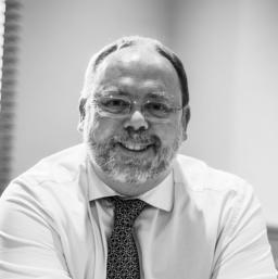 A grayscale image of an older man with a beard and glasses, smiling at the camera. He wears a light-colored dress shirt and a patterned tie. A window with blinds is partially visible in the background, giving an intimate glimpse into Iain Beveridge's sere